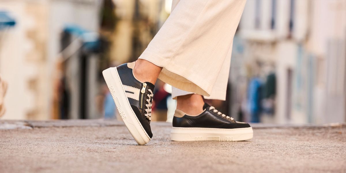 A person's feet and ankles as they walk outdoors, wearing black leather Rieker trainers with chunky cream platform soles, cream laces, and side zip detailing. They are wearing cropped cream trousers against a blurred street background.